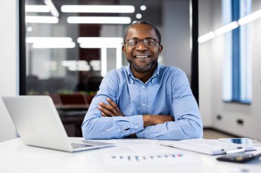 Ofis ortamında güvenle gülümseyen gözlüklü bir iş adamı. Masasında dizüstü bilgisayar ve belgelerle oturuyor, pozitiflik ve profesyonellik yayıyor..