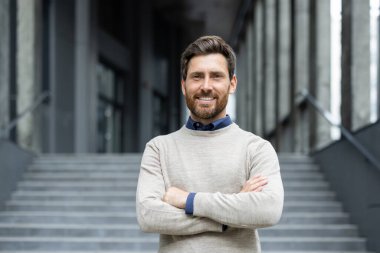 Modern ofis binasının dışında kolları çapraz duran kendine güvenen bir iş adamı. Günlük süveter giymek, profesyonel tavırlar sergilemek. Gülümseyen, rahatlatılmış ortam, ulaşılabilirliği iletir.