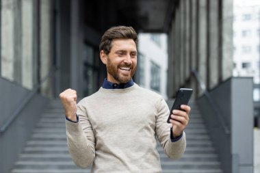 Sakallı, gülümseyen adam elinde telefonla olumlu haberi ya da başarıyı kutluyor. Kentsel ofis ortamında neşe ve başarıyı ifade etmek, heyecan ve mutluluk duygularını sergilemek.