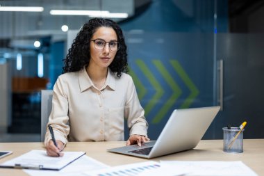 İspanyol iş kadını elinde kalemle dizüstü bilgisayarla masa başında çalışıyor. Modern ofis ortamında odaklanmayı ve analitik düşünmeyi gösterir, mesleki bağlılığı yansıtır