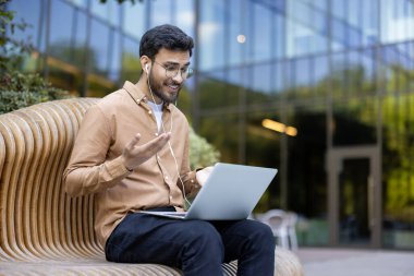 Kulaklık takan genç adam dışarıda tahta bankta oturuyor. Heyecan ve bağlantıyı gösteren bir video bağlantısı kuruyoruz. Modern yapı düzeni iş ve iletişimi geliştirir