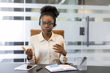 Ofiste video konferansında kulaklık takan kendine güvenen Afrikalı Amerikalı iş kadını. Verimli iletişim için jestler, tablet ve not defteri kullanan profesyonel tartışmalar