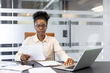 Profesyonel Afro-Amerikan iş kadını ofis masasında çalışıyor. Bilgisayarı açık olan belgeleri incelemeye odaklanmış. Kurumsal ortamı, odaklanmayı ve iş görevlerini temsil eder.