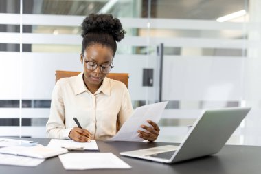 Profesyonel Afro-Amerikan iş kadını özveri ve odaklanma göstererek dizüstü bilgisayar ve belgelerle masa başında özenle çalışıyor. Görüntü modern ofis ayarlarını yansıtıyor