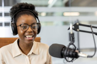 Afrika kökenli Amerikalı iş kadını, kulaklık takarak podcast sunuyor. Modern ofiste mikrofonla kayıt yapmakla meşgul. Profesyonel iletişimi ve yaratıcı bağlantıyı vurguluyor