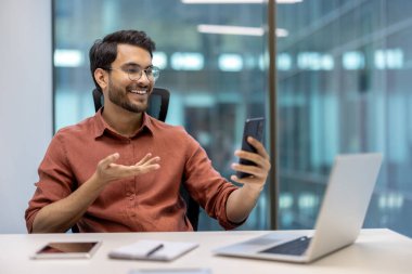 Modern ofisteki genç profesyonel akıllı telefon kullanarak görüntülü görüşme yapıyor. Mutlu ve kendinden emin, iş fırsatlarını tartışıyor. Masada dizüstü bilgisayar ve not defteriyle oturuyor.