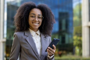 Modern ofis binasının dışında elinde telefonla gülümseyen Afro-Amerikan iş kadını. İş ortamında güven, iletişim ve teknolojiyi temsil eder