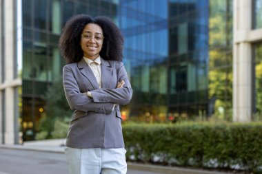 Ofis binasının dışında kolları bağlı duran kendinden emin Afrikalı Amerikalı iş kadını. Resmi iş kıyafetleri giyiyor. Güçlendirmeyi, liderliği ve profesyonelliği simgeliyor..