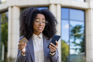 Afrikalı Amerikalı iş kadını, modern ofis binasının dışında kredi kartı ve akıllı telefon bulundurmaktan endişe duyduğunu söylüyor. Çevrimiçi bankacılık, mali yönetim ve dijital işlemleri temsil eder.