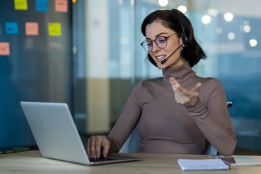 Video görüşmesi sırasında dizüstü bilgisayar kullanan kendine güvenen profesyonel bir kadın. Kulaklık takmak, modern ofis ortamında önemli iş konularını tartışmak. Not defteri ve telefon referans için masaya..