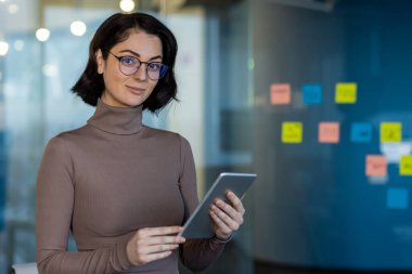 Modern ofiste tablet tutan kendine güvenen bir iş kadını. Renkli yapışkan notların yanında duruyor. Üretkenlik, teknoloji, güven ve ofis işi gibi kavramlar için ideal.