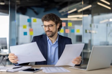 Ofis masasındaki kağıtları analiz eden kendine güvenen bir işadamı. Bilgisayarlı ve telefonlu profesyonel ayarlar, verimliliği arttırıyor. Modern çalışma alanı renkli yapışkan notlar, yaratıcı düşünce için ideal