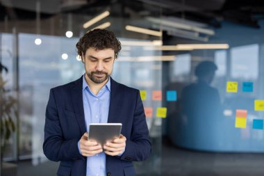 Resmi giyinmiş iş adamı, çağdaş ofis ortamında stratejik planlama tabletine odaklandı. Cam duvardaki yapışkan notlar beyin fırtınası sürecini yansıtıyor.