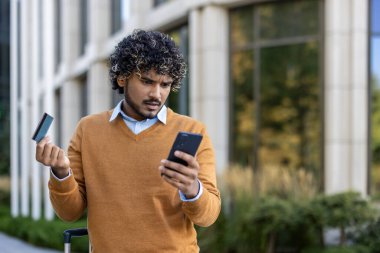 Kıvırcık saçlı, kahverengi kazaklı kredi kartı ve akıllı telefon tutan bir adam. Dışarıda dikilmiş, kafası karışmış, muhtemelen iş gezisi sırasında internet bankacılığı ya da ödeme sorunuyla karşı karşıya..