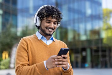 Şehir ortamında akıllı telefon ve kulaklık kullanan gülümseyen genç bir adam. Görüntü gündelik yaşam tarzı, teknoloji etkileşimi ve modern iletişimi yakalar.