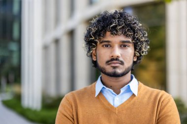 Kıvırcık saçlı, süveter ve gömlek giyen kendinden emin bir gencin portresi. Şehir ortamında dışarıda durmuş, ciddi bir ifadeyle kameraya bakıyor. Profesyonel havayı yakalar.