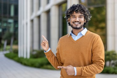 Modern ofis binasının önünde duran gülümseyen genç profesyonel iki elini de rahatça işaret ediyor. Resim, iş veya akademik çevrede olumluluk, başarı ve coşku taşır.