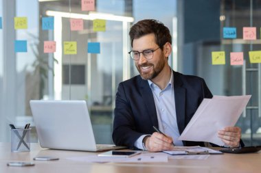 Businessman in suit engaging in office work with documents, laptop, and smartphone. Smiling professional demonstrating productivity and focus, seated at desk in modern workspace