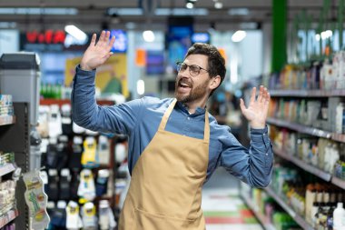 Önlük giyen neşeli bir erkek market çalışanı koridorda çalışmaktan zevk alıyor ve heyecan duyuyor. Etrafı çeşitli ürünlerle dolu raflarla çevrili, canlı ve canlı bir atmosfer yaratıyor.