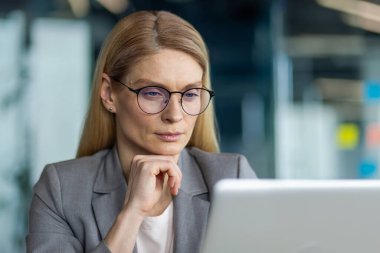 Ofiste dizüstü bilgisayarla çalışan, gözlüklü kendine güvenen bir iş kadını. İfade odaklanmayı, düşünmeyi ve kararlılığı gösterir. Modern ofis ayarları profesyonel atmosferi geliştirir.