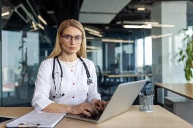 Gözlüklü profesyonel kadın doktor modern ofis ortamında dizüstü bilgisayarla çalışıyor. Steteskop ve yakınlardaki belgeleri içeren tıbbi görevlere odaklanmış.