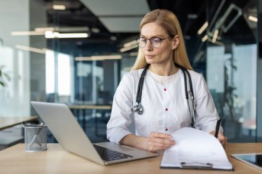 Professional female doctor in white coat with stethoscope working on laptop and examining medical documents. Scene conveys focus, expertise, and concentration in healthcare and medical field. clipart