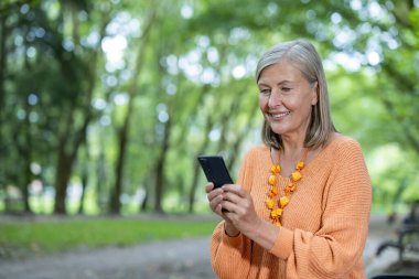 Yeşillik bir park ortamında akıllı telefon kullanan son sınıf öğrencisi. Sahne, boş zaman, teknoloji kullanımı ve doğa ile bağlantı hissini iletir.
