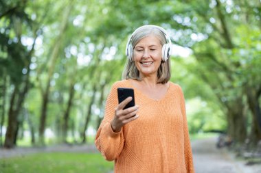 Turuncu kazaklı yaşlı bir kadın parkta kulaklık takıyor, keyifle gülümsüyor. Boş zaman ve teknolojiyi kucaklamak, aktif yaşam tarzı ve bağlantıyı sembolize etmek.