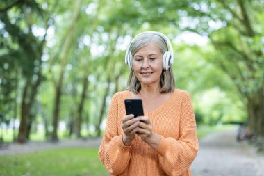 Kulaklık takan yaşlı bir kadın, park ortamında telefon kullanıyor. Mutlu görünüyor, teknolojiyle meşgul, müzikten hoşlanıyor. Eski nesiller arasındaki bağlantı ve teknoloji kullanımını temsil eder