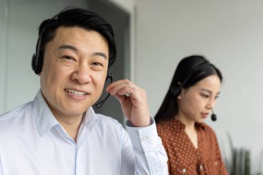 Asyalı iş adamları iş birliği yapıyor. Gülümseyen adam ve odaklanmış kadın kulaklık takıyor, profesyonel müşteri desteği alıyor. Takım çalışması, iletişim ve müşteri hizmetlerini gösterir