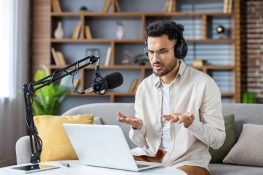 İspanyol adam evde podcast kaydı için dizüstü bilgisayar ve mikrofon kullanıyor. Kulaklık takmak, mikrofona konuşmak, çevrimiçi içerik yaratmak. Modern teknoloji kurulumu telefon ve kayıt ekipmanlarını içerir.