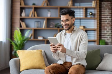 Gözlüklü ve gömlekli genç bir Hintli evdeki kanepede oturur ve gülümsemeli bir tablet kullanır..