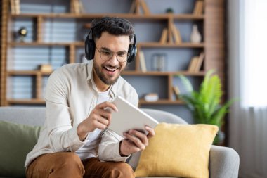 Oyun oynamak için tablet kullanan kulaklık takan gülümseyen genç adam. Oturma odasında rahat bir atmosfer, gündelik giysiler, modern ev ortamında keyif ve eğlence göstergesi..