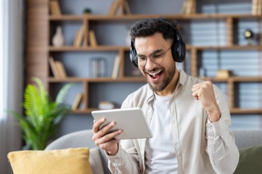 Kulaklık takan adam elinde tablet tutarken başarıyı coşkuyla kutluyor. Modern oturma odasında oturmuş, neşe ve başarıyı ifade ediyor..