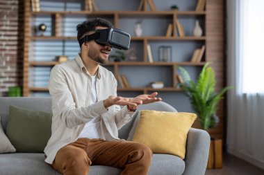 Genç adam VR kulaklık takarak kanepeye oturur ve modern oturma odasında sanal gerçekliği tecrübe eder. Elleriyle dijital dünyada jestler yapıyor, evdeki teknoloji ve yenilikten zevk alıyor..