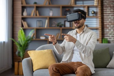 Young man immersed in virtual reality using VR headset, sitting on sofa and interacting with virtual environment. Smiling enthusiastically, experiencing cutting-edge technology excitement