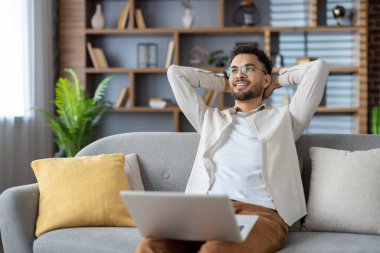 Kanepede dizüstü bilgisayarla oturan gülümseyen adam evde rahat bir anın tadını çıkarıyor. Günlük kıyafetler, eller başının arkasında, rahatlık ve memnuniyeti ifade ediyor. Kitaplıklarla modern oturma odası ayarları