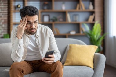 Adam evde akıllı telefon kullanarak hayal kırıklığı yaşıyor, kanepede oturuyor, alnında ellerle stres gösteriyor. Günlük kıyafetler rahatlatıcı ama endişeli atmosfere katkı sağlar..