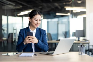 Ofiste çalışan Asyalı iş kadını akıllı telefon ve dizüstü bilgisayar kullanıyor. Profesyonel kıyafet, iş görevlerine odaklanmış. Şirket ortamında modern teknoloji kullanımını temsil eder.