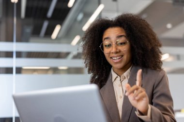 Takım elbiseli ve gözlüklü Afro-Amerikalı genç iş kadını ofis masasında oturuyor ve bilgisayardaki video görüşmesinde konuşuyor..