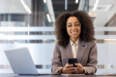 Takım elbiseli, kıvırcık saçlı, elinde cep telefonu olan ve kameraya bakan gülümseyen Afro-Amerikan bir kadının portresi..