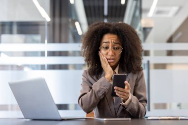 Şok olmuş ve korkmuş genç Afrikalı Amerikalı kadın ofiste, masada oturuyor ve cep telefonu ekranına bakıyor, elini yanağına doğru tutuyor..