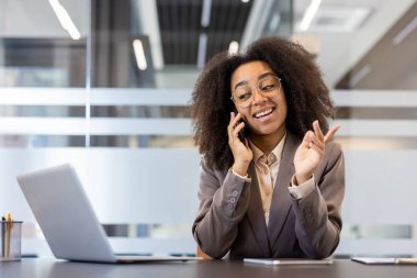 Gülümseyen ve başarılı genç Afrikalı Amerikalı iş kadını masadaki ofiste oturuyor ve elleriyle el kol hareketi yaparken telefonla konuşuyor..