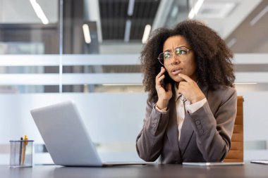 Ciddi bir Afro-Amerikalı kadın takım elbise içinde ofiste oturuyor ve telefonla konuşuyor, problemleri ve soruları çözüyor..