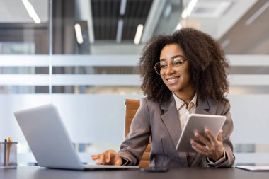 Önlüklü ve takım elbiseli gülümseyen Afro-Amerikalı bir kadın ofiste dizüstü bilgisayarda çalışıyor ve tablet kullanıyor..