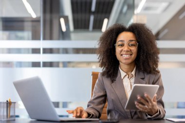 Ofiste çalışan gülümseyen ve başarılı bir Afrikalı Amerikalı iş kadınının portresi. Bir dizüstü bilgisayar ve tablet kullanarak kameraya güvenle bakıyor..