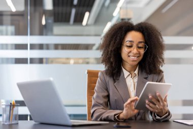 Takım elbiseli ve gözlüklü Afro-Amerikalı genç bir iş kadını ofisteki bir masada oturuyor ve gülümseyerek tablet kullanıyor..