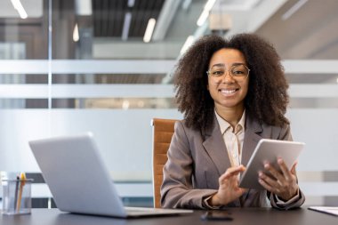 Takım elbiseli ve önlüklü gülümseyen kıvırcık saçlı genç bir kadının portresi bir masada elinde bir tabletle oturup kameraya bakıyor..