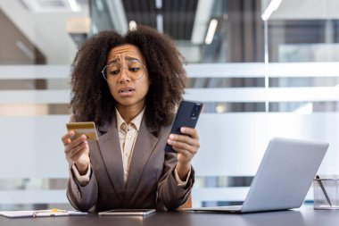 Ofiste oturan, elinde kredi kartıyla cep telefonu ekranından hayal kırıklığına uğramış, üzgün, takım elbiseli Afro-Amerikan bir kadın..