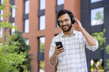 Memnun ve gülümseyen genç Hintli bir adam şehir sokaklarında kulaklık takıyor ve cep telefonu kullanıyor..